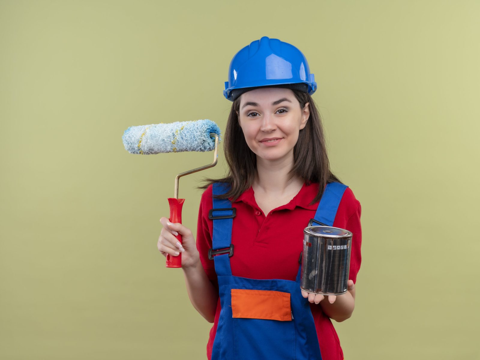 smiling young builder girl with blue safety helmet holds paint roller and paint on isolated green background